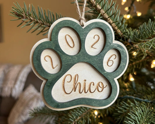 A green wooden ornament shaped like a paw print with the name "Chico" engraved on it, hanging on a Christmas tree.