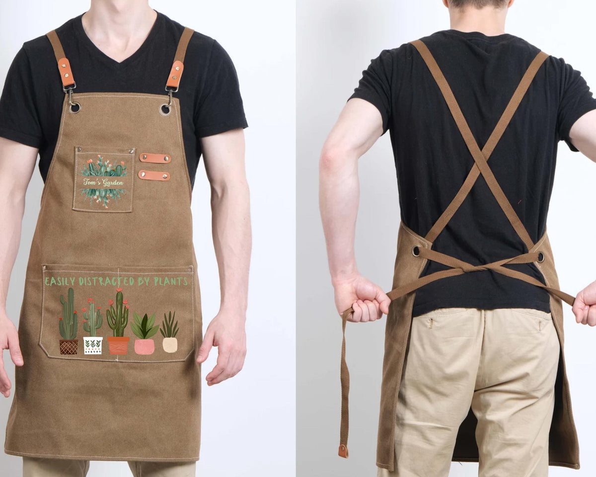 a man wearing a tan apron with a design of cacti and plants. The apron has a pocket on the front and straps across the back.