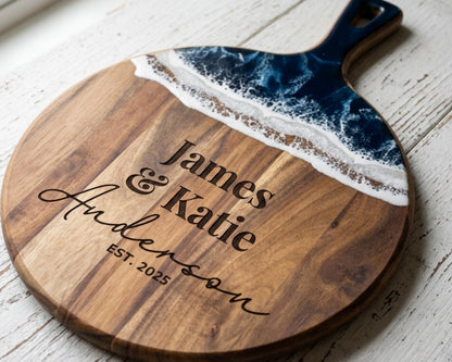 A wooden cutting board with a blue and white wave design, engraved with the names "James & Katie Anderson" and the establishment year "EST. 2025".