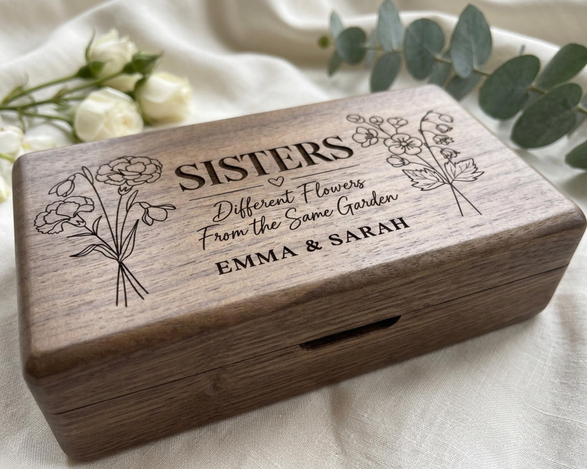 A wooden box with the text "SISTERS Different Flowers From the Same Garden" engraved on it, along with illustrations of flowers. The box is placed on a white cloth, and there are some flowers in the background.