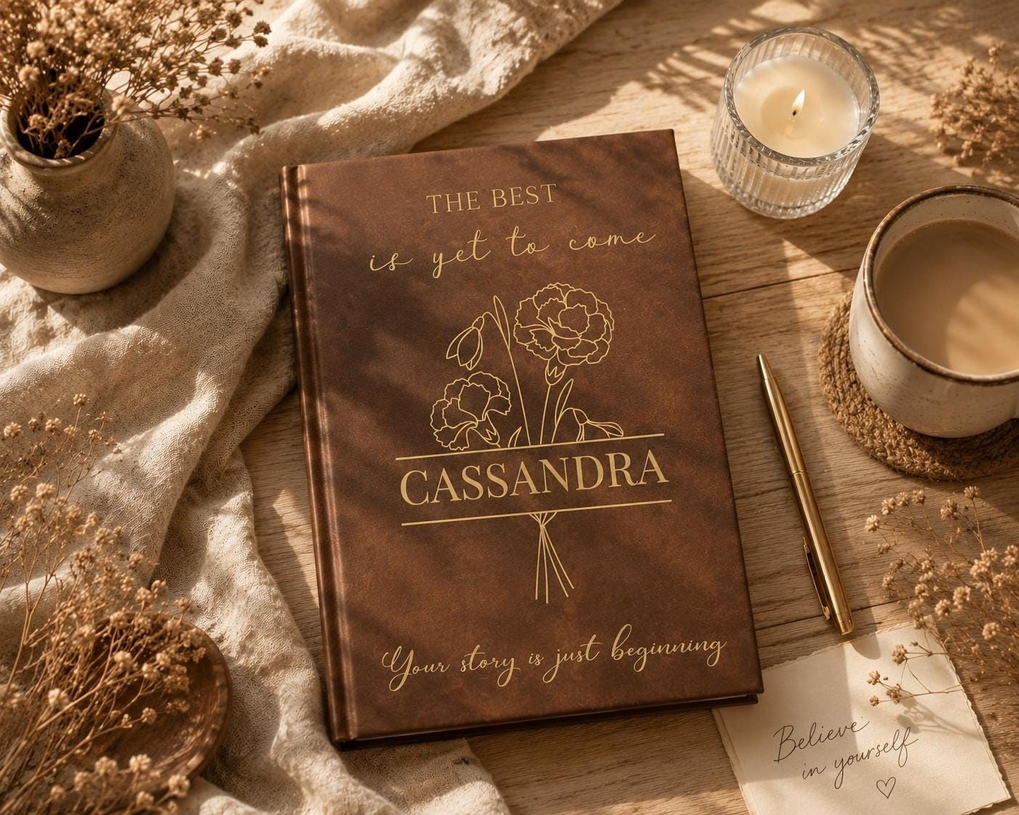 a brown notebook with the title "CASSANDRA" and the phrase "Your story is just beginning" written on it, along with a pen and a candle. The notebook is placed on a wooden surface, surrounded by various objects such as a vase with flowers, a cup, and a note.