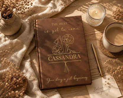 a brown notebook with the title "CASSANDRA" and the phrase "Your story is just beginning" written on it, along with a pen and a candle. The notebook is placed on a wooden surface, surrounded by various objects such as a vase with flowers, a cup, and a note.
