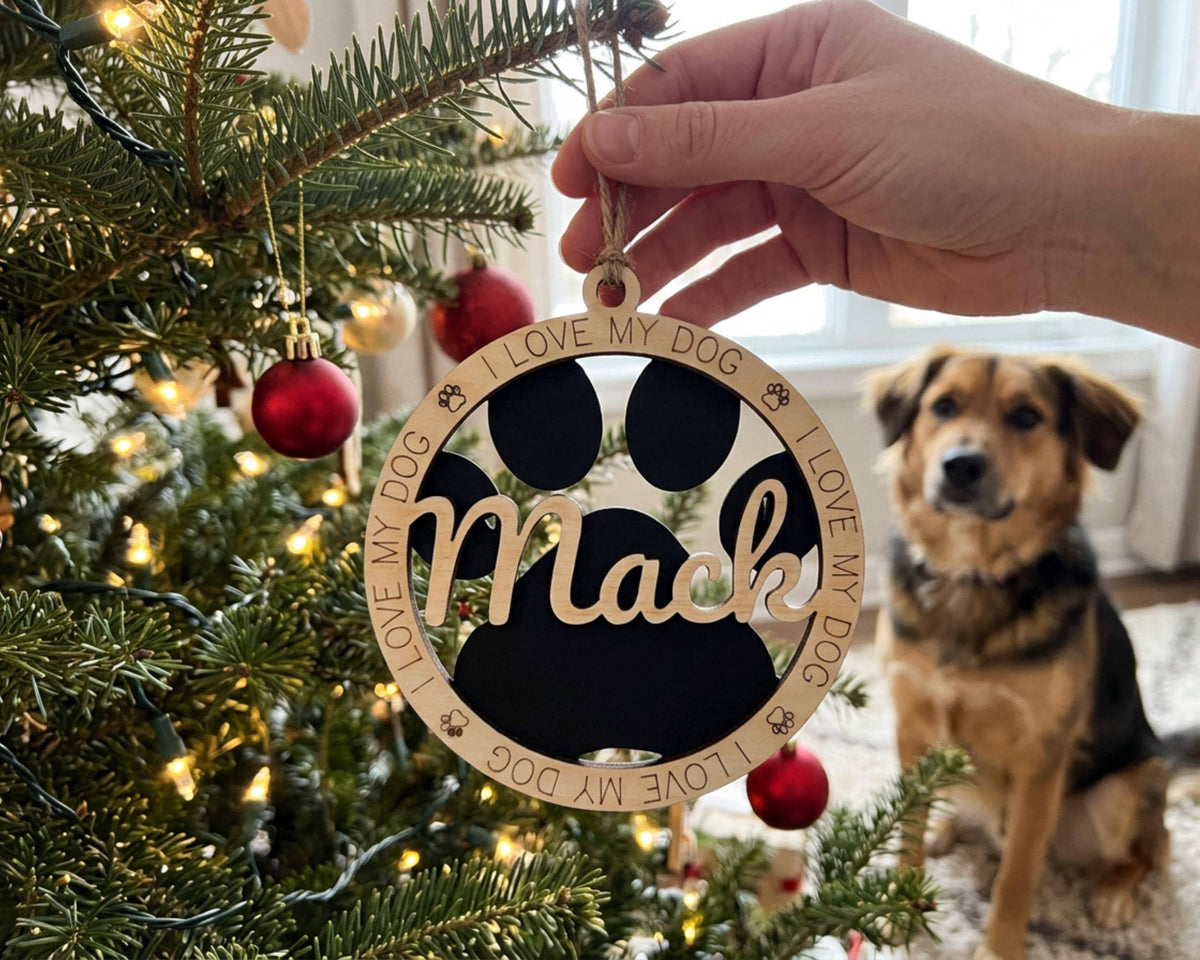 A hand is holding a wooden ornament with the name "Mac" on it, hanging from a Christmas tree. A dog is sitting in the background, looking at the ornament.