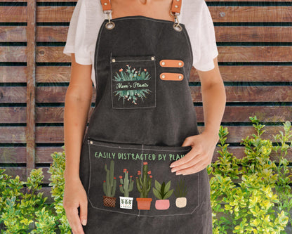 A person wearing a black apron with a plant design and text printed on it, standing in front of a wooden fence with greenery.