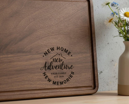 A wooden cutting board with a decorative design and text that says "NEW HOME. NEW ADVENTURE. RIKER FAMILY. EST. 2028" is placed next to a vase with flowers on a table.