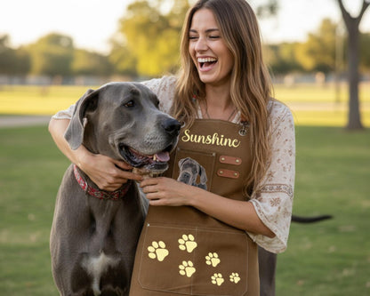 A woman in a brown apron with a dog in a park, both smiling and enjoying each other's company.