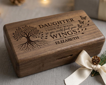 A wooden box with a tree design and text engraved on it, placed on a table with a pine cone and a ribbon.