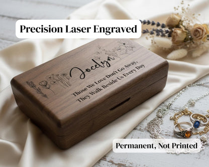 A wooden box with a personalized engraved message, placed on a white cloth, with a bouquet of flowers and jewelry in the background.