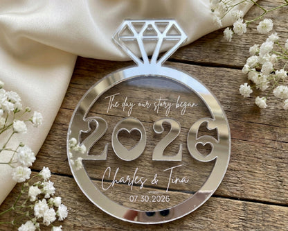 A clear glass ornament with the numbers "2026" and the names "Charles & Tina" written on it, placed on a wooden surface with white flowers.
