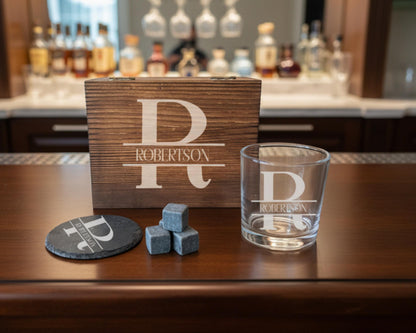 a wooden box with a personalized monogrammed name on it, a glass with a personalized monogrammed name, and some gray rocks on a wooden surface.