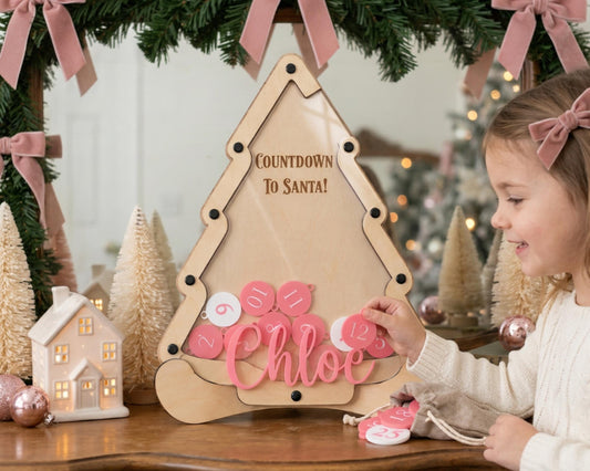 A young girl is playing with a pink and white countdown timer shaped like a Christmas tree, surrounded by holiday decorations and ornaments.