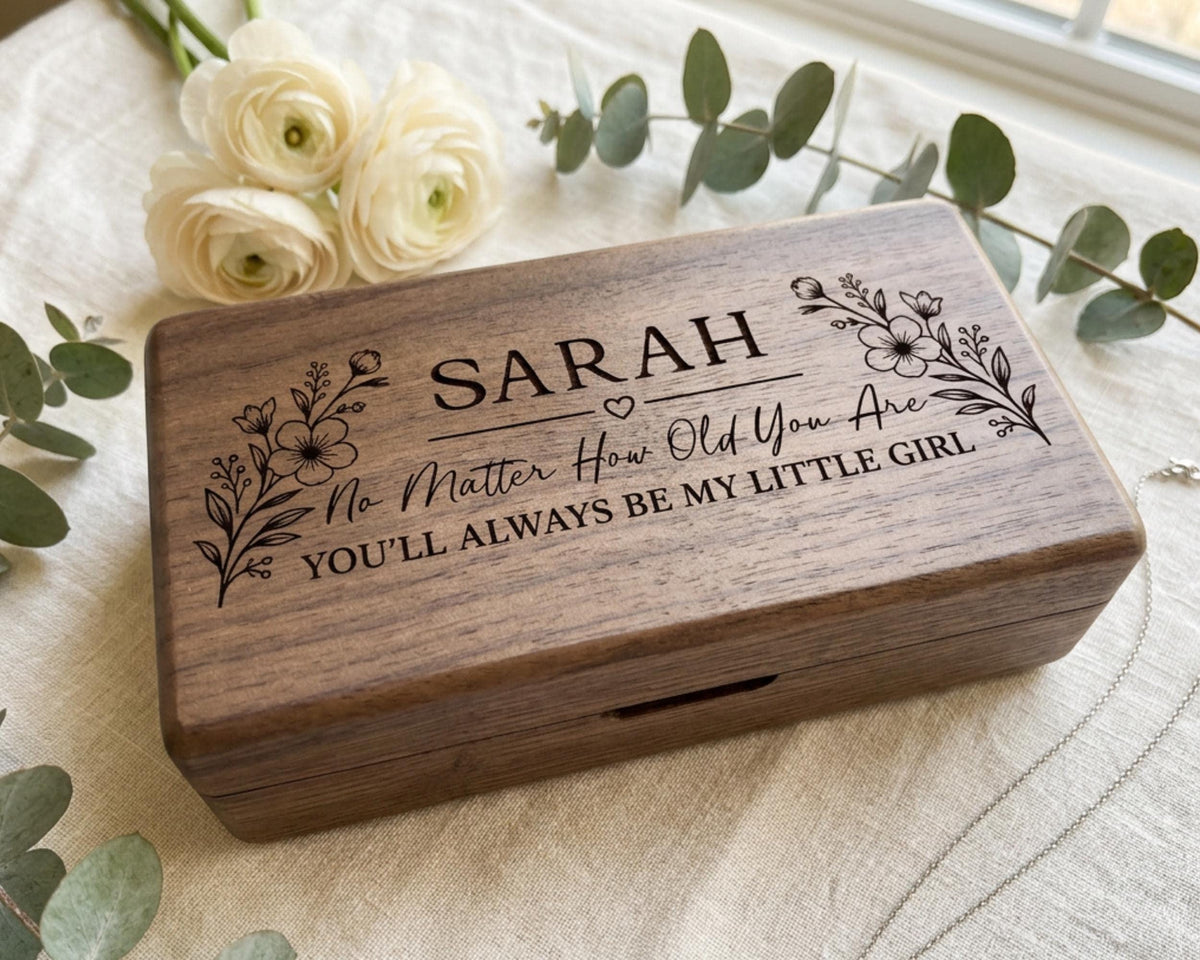 A wooden box with the name "SARAH" engraved on it, surrounded by white flowers and greenery.