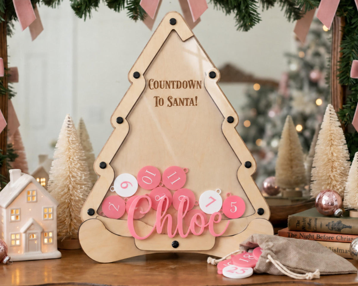 A wooden countdown clock with the text "Countdown To Santa!" and the name "Chloe" written on it, surrounded by pink and white ornaments and a small Christmas tree.