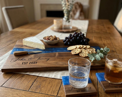 a wooden cutting board with a variety of food items, including cheese, grapes, crackers, and a knife, placed on a dining table. There are also two glasses of water and two glasses of tea or other beverages.