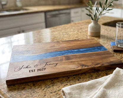 A wooden cutting board with a blue river-like design on it, placed on a kitchen counter. The cutting board has the names "John & Tommy" engraved on it, and there is a vase with greenery and a glass of water nearby.