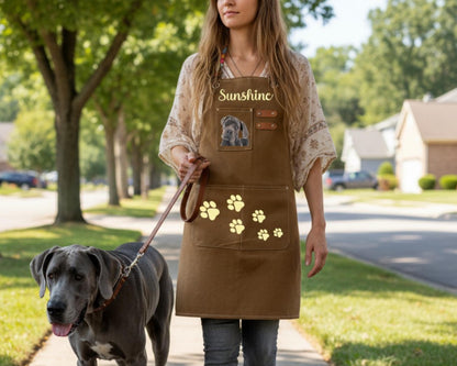 A woman wearing a brown apron with a dog walking on a leash is walking down a sidewalk.