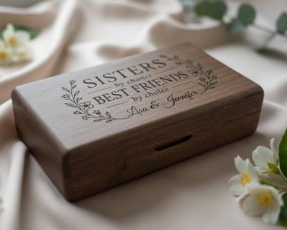 A wooden box with the text "SISTERS by chance by best friends by choice" engraved on it, along with the names "Anna & Jennifer" below. The box is placed on a fabric surface, with some white flowers visible in the background.