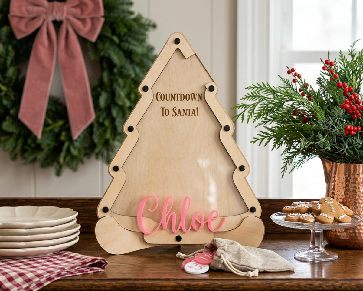 A wooden Christmas tree-shaped countdown timer with the name "Chloe" written on it, placed on a wooden table. The table also has a plate of cookies, a wreath, and a vase of red berries.