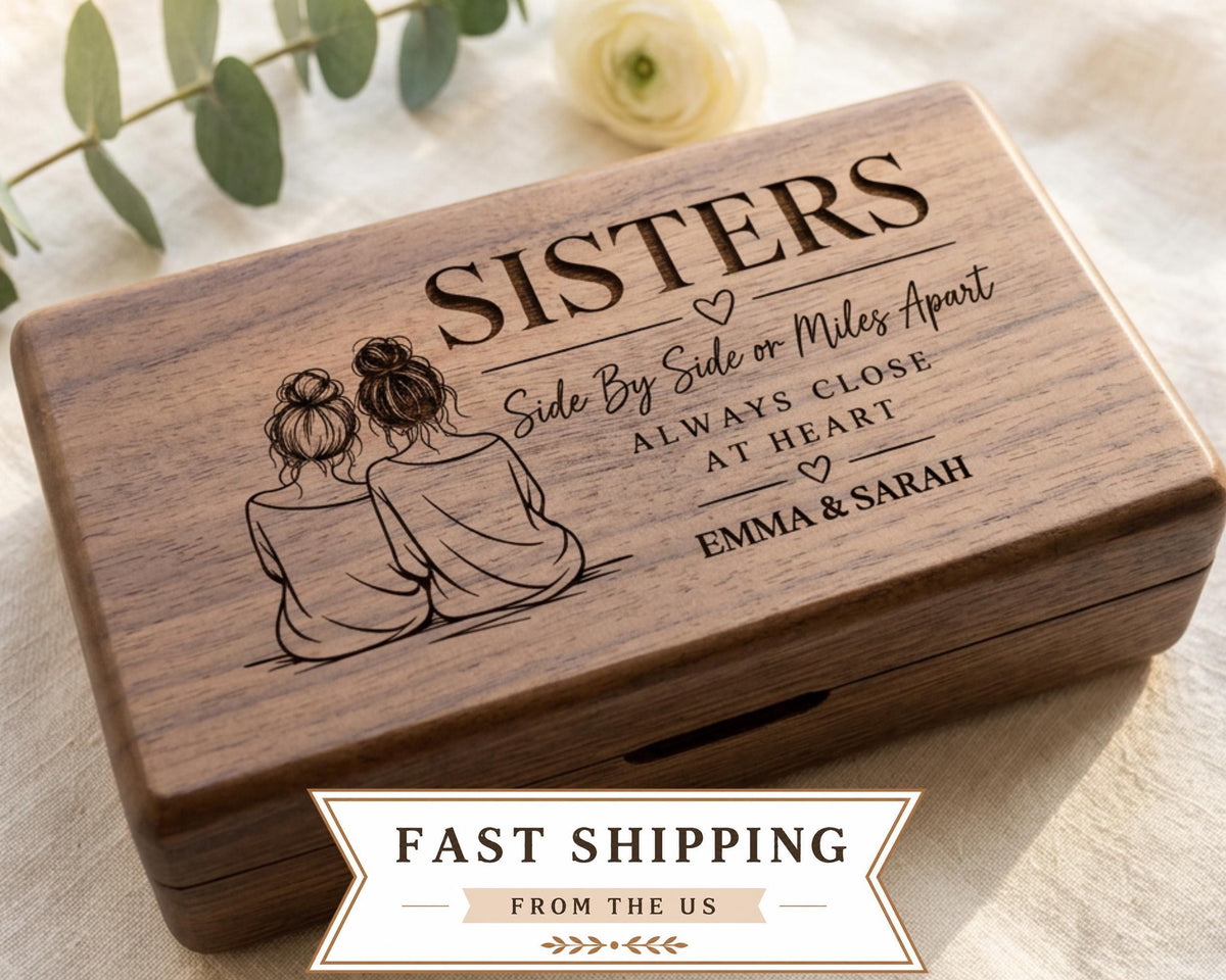 A wooden box with a design featuring two women sitting next to each other, along with the text "SISTERS Side By Side or Miles Apart Always Close at Heart Emma & Sarah". The box is placed on a white cloth, and there is a rose in the background.