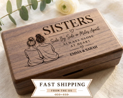 A wooden box with a design featuring two women sitting next to each other, along with the text "SISTERS Side By Side or Miles Apart Always Close at Heart Emma & Sarah". The box is placed on a white cloth, and there is a rose in the background.
