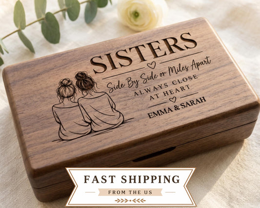 A wooden box with a design featuring two women sitting next to each other, along with the text "SISTERS Side By Side or Miles Apart Always Close at Heart Emma & Sarah". The box is placed on a white cloth, and there is a rose in the background.