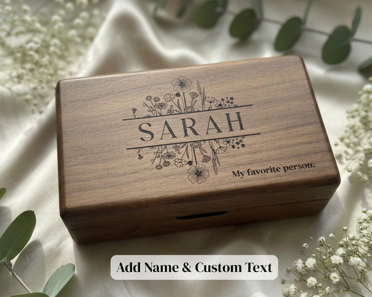 A wooden box with the name "SARAH" engraved on it, surrounded by flowers and greenery.