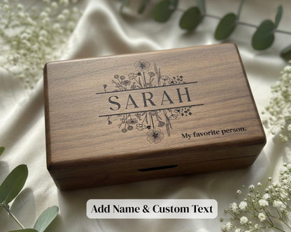 A wooden box with the name "SARAH" engraved on it, surrounded by flowers and greenery.