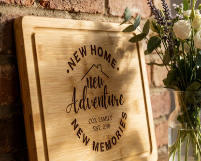 A wooden cutting board with the text "NEW HOME. new Adventure. COX FAMILY. new Memories" engraved on it, along with a vase of white flowers and greenery.