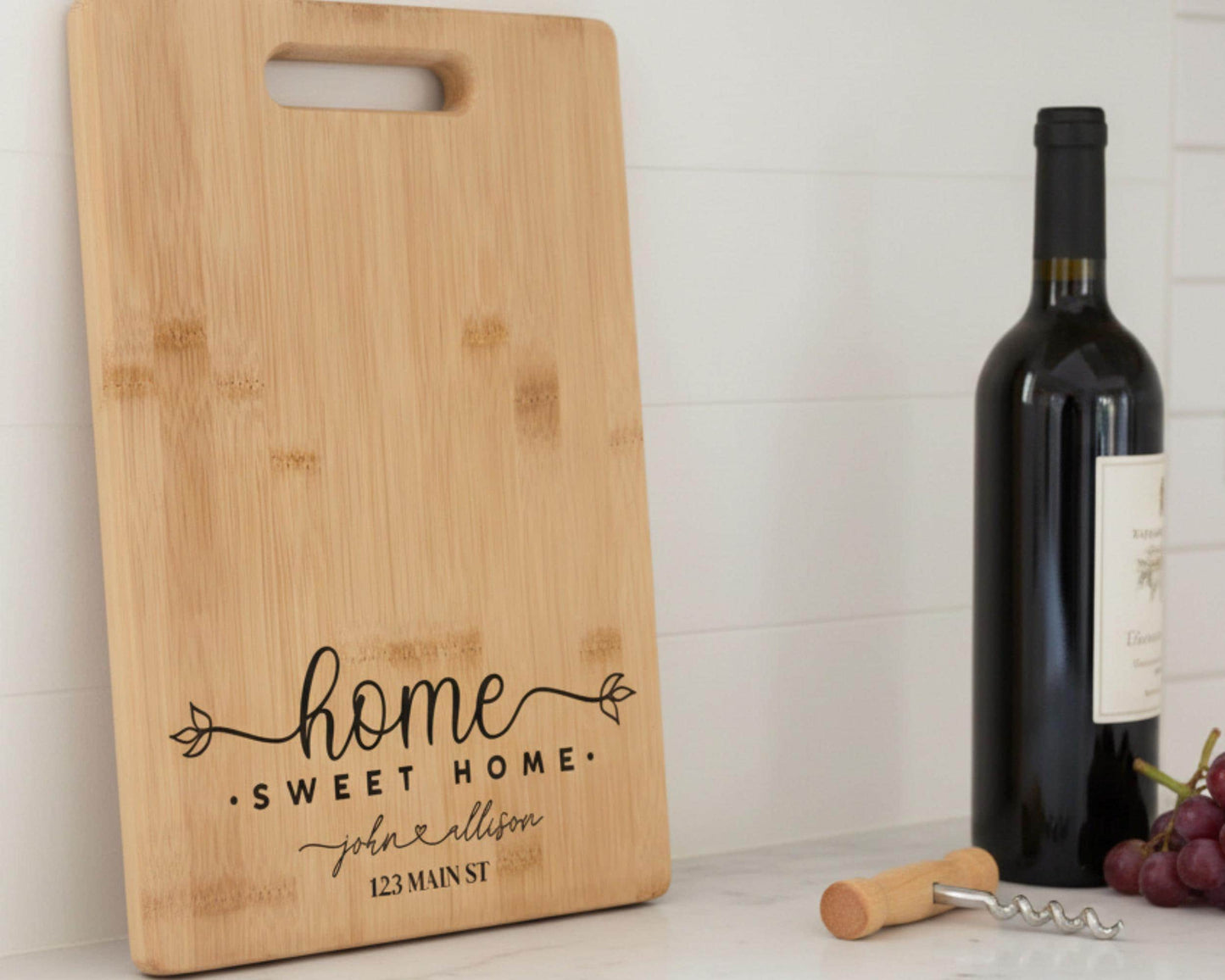 a wooden cutting board with the text "home sweet home" and "John Allison" printed on it, along with the address "123 Main St." Next to the cutting board is a bottle of wine and a corkscrew, suggesting a home-themed setting.