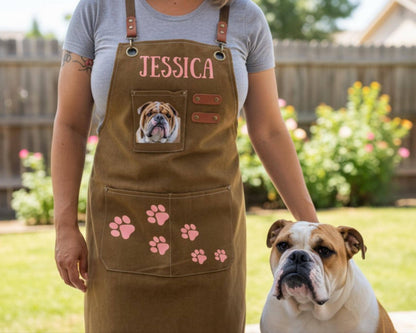 A woman wearing a brown apron with a dog's face and paw prints on it stands next to a brown and white bulldog in a garden setting.