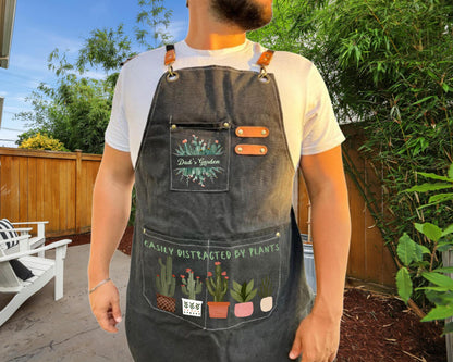 A man wearing a gray apron with a plant design stands in front of a wooden fence and greenery.