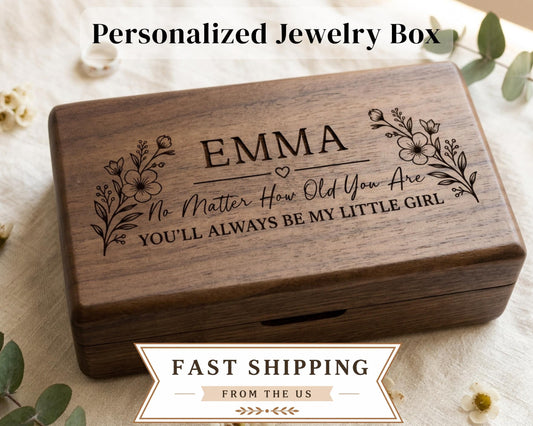 A wooden jewelry box with the name "Emma" engraved on it, surrounded by floral designs. The box appears to be personalized and is placed on a white cloth with greenery.