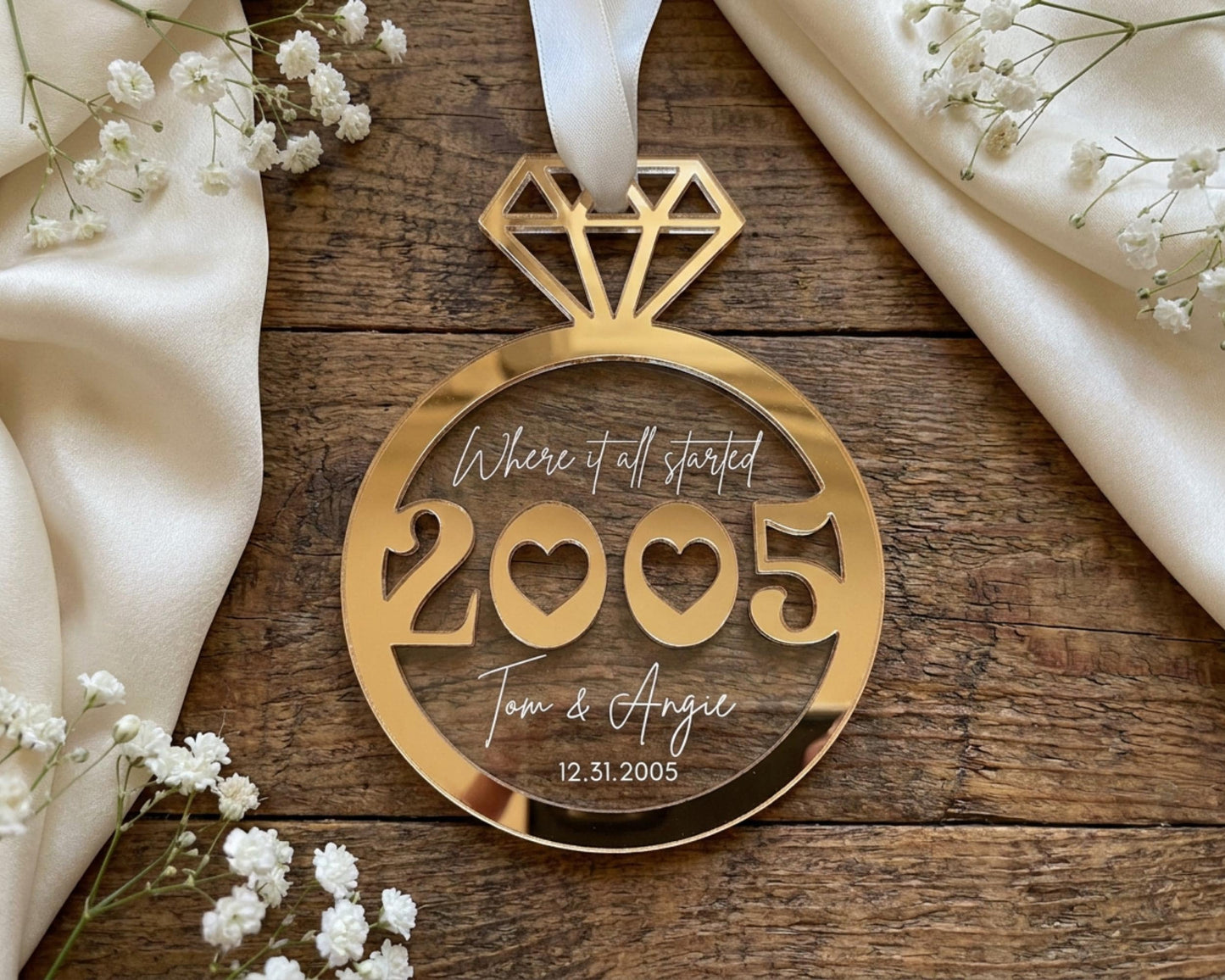 A gold-colored ornament with the text "Where it all started 2005 Tom & Angie 12.5.2005" is displayed on a wooden surface, surrounded by white flowers.