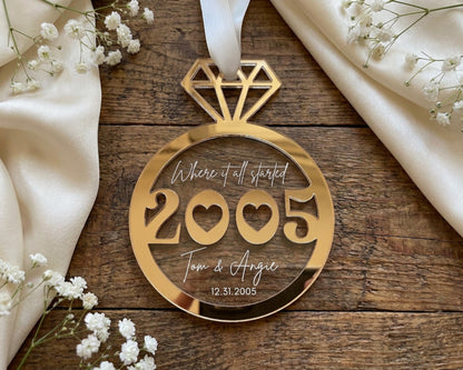 A gold-colored ornament with the text "Where it all started 2005 Tom & Angie 12.5.2005" is displayed on a wooden surface, surrounded by white flowers.