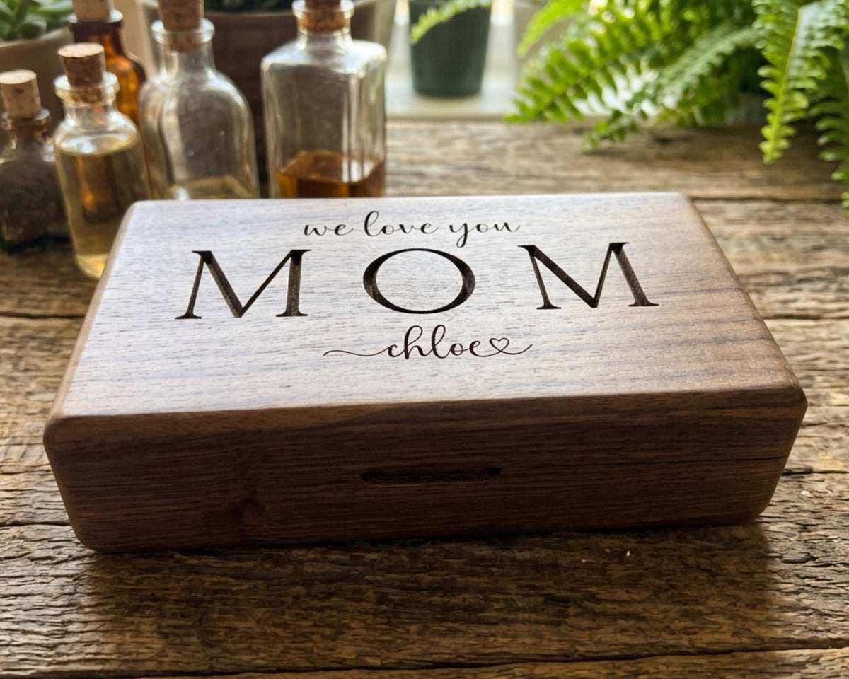 A wooden box with the text "we love you Mom" engraved on it, placed on a wooden surface with various bottles and plants in the background.