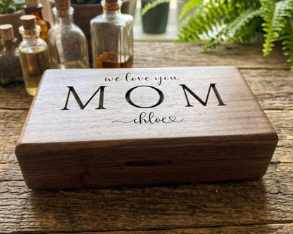 A wooden box with the text "we love you Mom" engraved on it, placed on a wooden surface with various bottles and plants in the background.