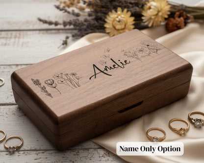 A wooden box with the name "Alicie" engraved on it, surrounded by various rings and flowers on a wooden surface.
