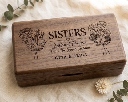 A wooden box with the text "SISTERS Different Flowers From the Same Garden GINA & ERICA" engraved on it, along with illustrations of flowers. The box is placed on a white cloth, and there are some flowers and greenery in the background.
