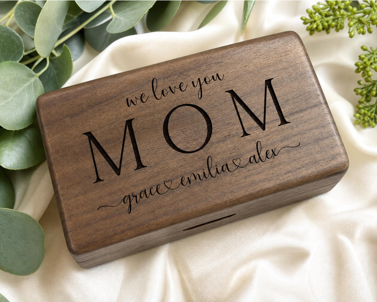 A wooden box with the inscription "we love you Mom Grace + Alex" engraved on it, placed on a white cloth with greenery.