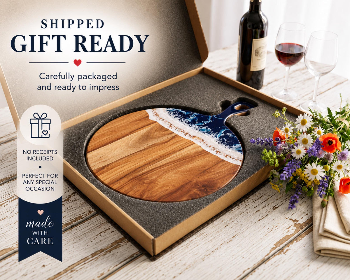 a wooden cutting board with a blue and white design, placed inside a cardboard box on a wooden table. There are also wine glasses and a bouquet of flowers nearby.