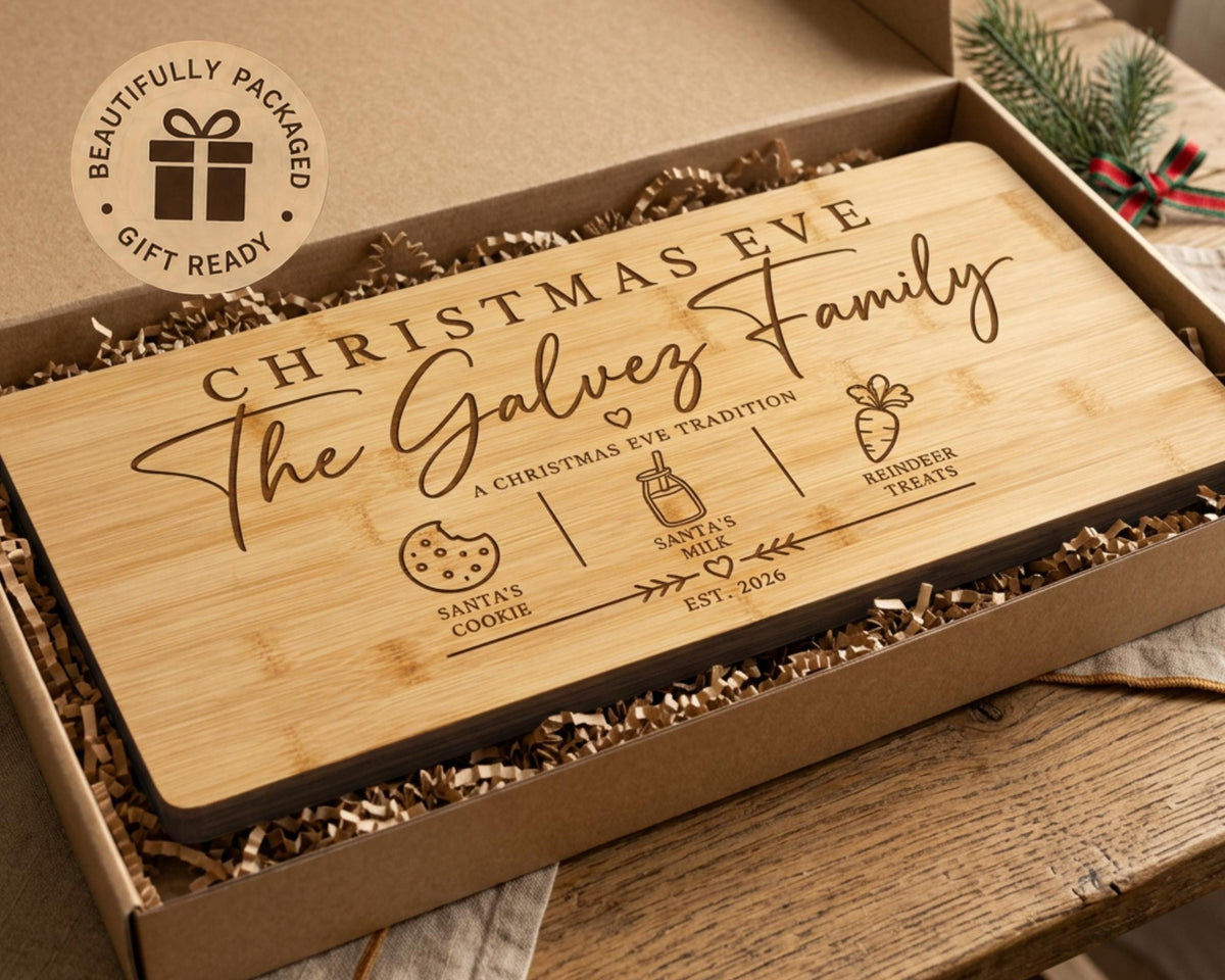 A wooden cutting board with the text "Christmas Eve The Galviz Family" engraved on it, along with icons representing Santa's cookie, milk, and treats. The cutting board is placed in a cardboard box with a gift tag that reads "Beautifully packaged Gift Ready".