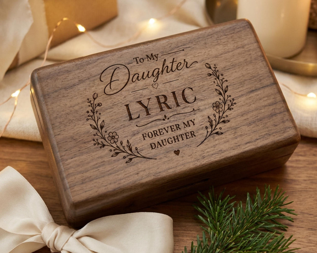 A wooden box with a decorative design and text engraved on it, placed on a wooden surface with a ribbon and greenery in the background.