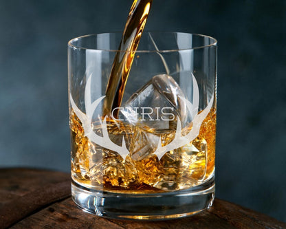 A glass of amber-colored liquid, likely whiskey, with ice cubes and a golden liquid being poured into it. The glass has a personalized name etched on it.