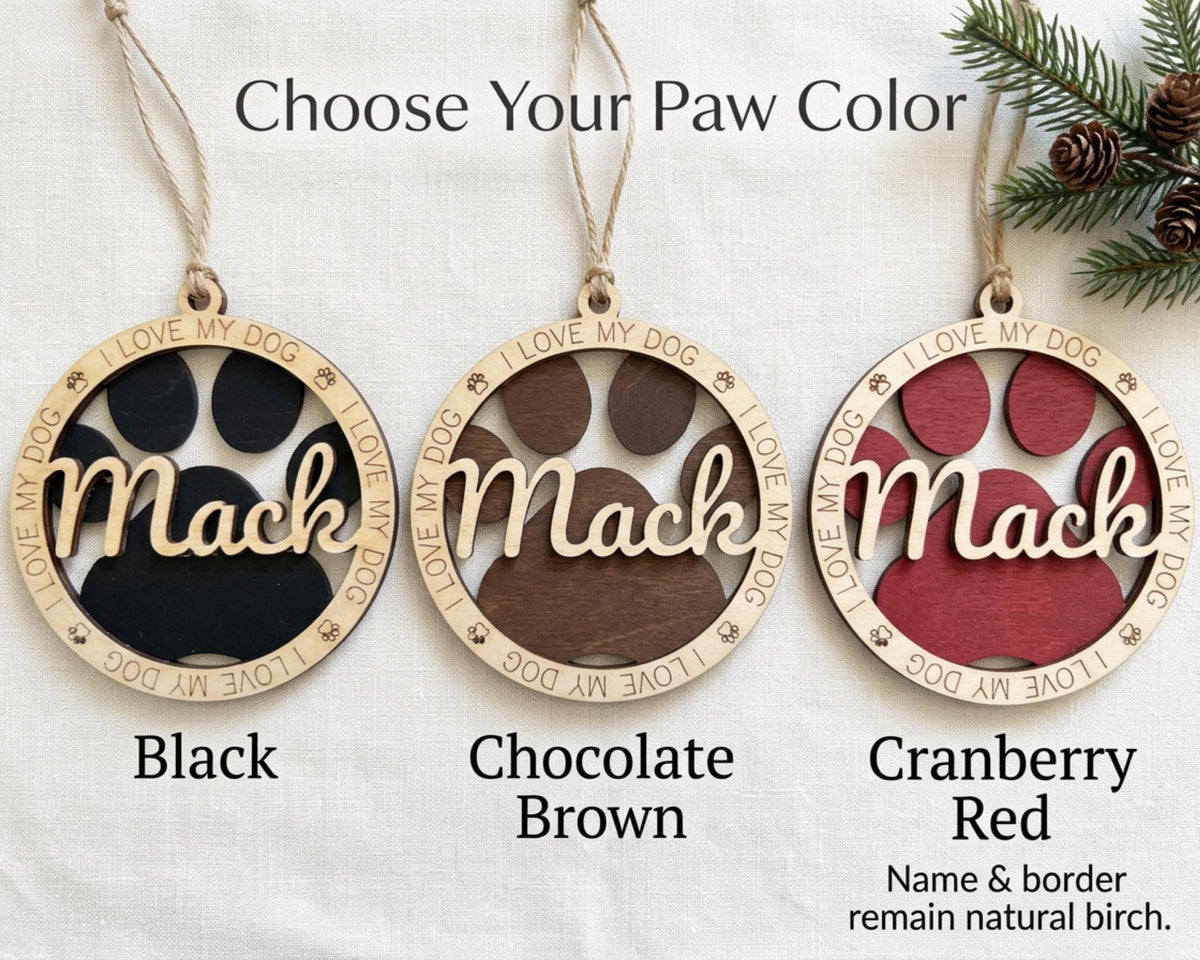 three circular ornaments with the name "Mac" written on them, each in a different color: black, chocolate brown, and cranberry red. The ornaments are hanging from strings, and there is a pine branch visible in the background.
