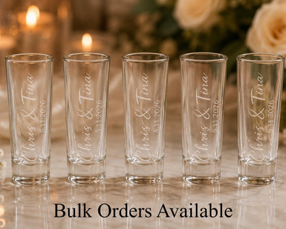 A row of five shot glasses with personalized names and dates engraved on them, placed on a table with a floral arrangement in the background.