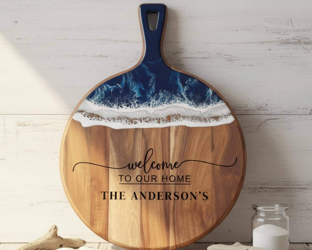 A wooden cutting board with a blue handle and a wave-like design on the top surface, accompanied by the text "welcome to our home THE ANDERSON'S" on the front.