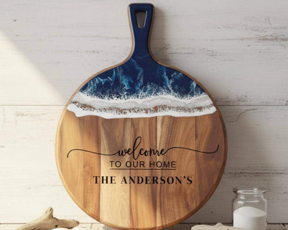 A wooden cutting board with a blue handle and a wave-like design on the top surface, accompanied by the text "welcome to our home THE ANDERSON'S" on the front.
