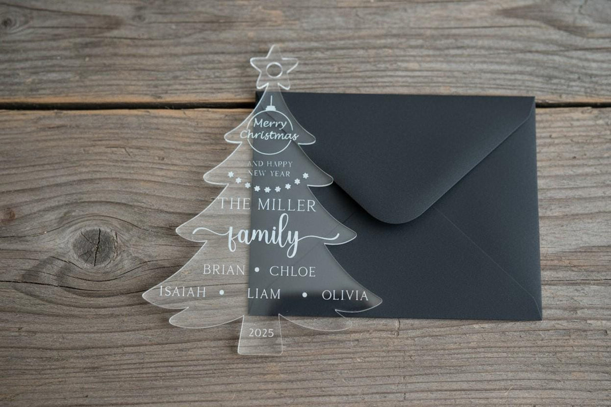 Clear acrylic Christmas tree card with engraved family names resting on a wooden table next to a black envelope