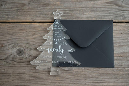 Clear acrylic Christmas tree card with engraved family names resting on a wooden table next to a black envelope