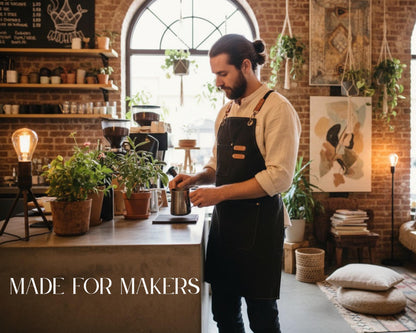 A bearded man in a black apron is preparing drinks at a counter in a cozy, rustic-looking coffee shop, surrounded by potted plants and other decor.